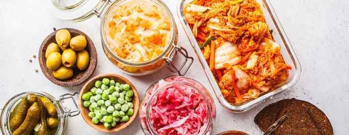 Assortment of fermented and pickled foods, including kimchi, sauerkraut, pickled onions, olives, cucumbers, and peas, arranged on a white background.